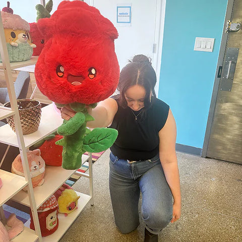 Image shows a woman kneeling and holding out the stuffed red rose. The petals are larger than her head.