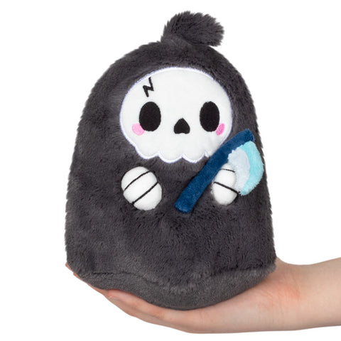 A plush toy in the shape of a stylized reaper with a skull face, holding a blue scythe. It is held in someone's hand.