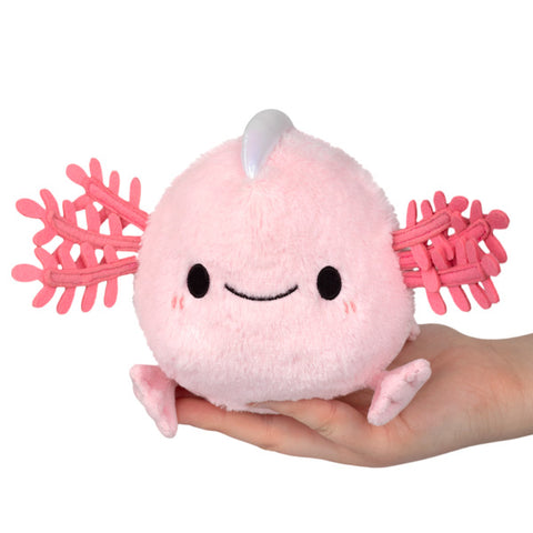 A plush toy resembling a baby axolotl with a pink body and gills, being held in a person's hand.