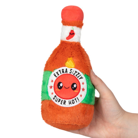 A plush toy shaped like a bottle of hot sauce with a cute face on the label, held in a person's hand.