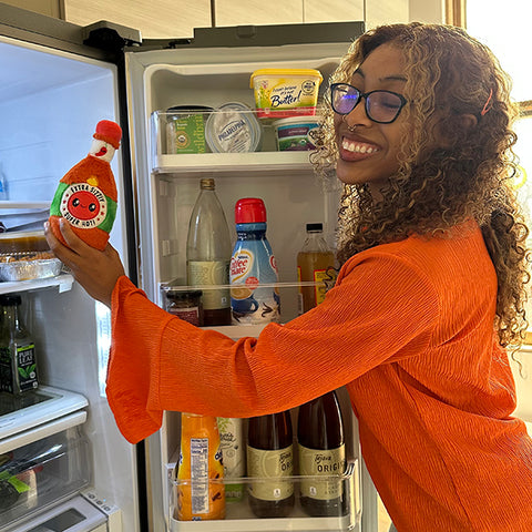 A woman holds the plush hot sauce near a fridge. It is slightly smaller than her head.