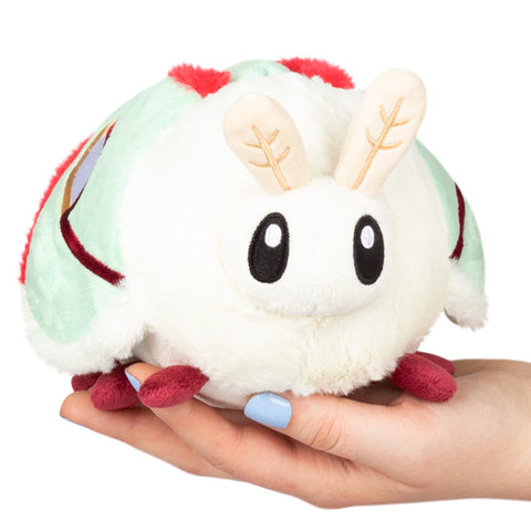 A plush toy resembling a Luna moth with white body, red and green accents, and large black eyes, being held in a person's hand.