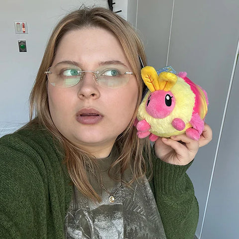 Image shows a woman holding the stuffed rosy maple moth up to her face. It is about half the size of her head.