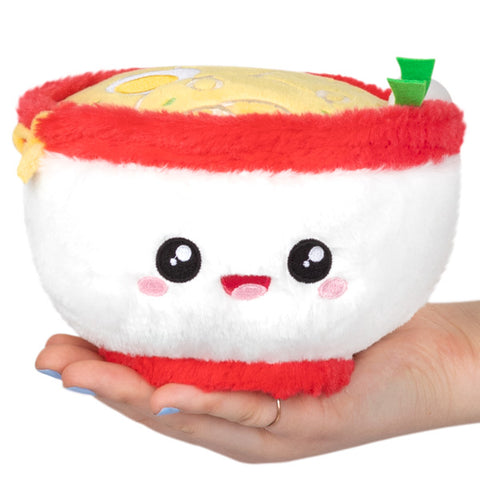 A plush toy resembling a bowl of ramen with a happy face, holding in a human hand.