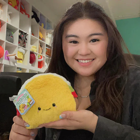 A woman holds the plush taco near her face. It is slightly smaller than her head.