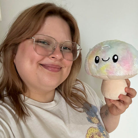 Image shows a woman holding the stuffed tie dye mushroom up to her face. It is slightly smaller than her head.