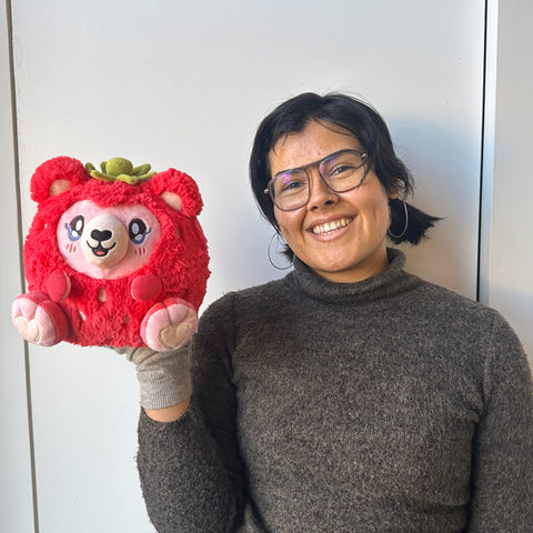 A woman holds the plush strawberry bear near her face. It is slightly larger than her head.