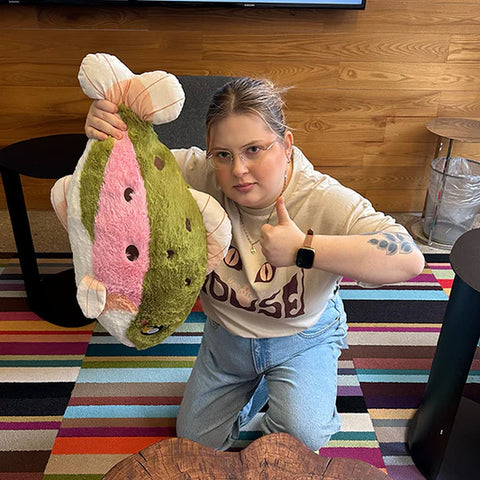 Image shows a woman holding the stuffed rainbow trout up by the tail. It is approximately the length of her torso,