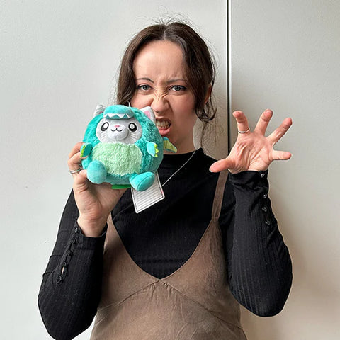 A woman holds the plush dragon cat near her face. It is slightly smaller than her head.