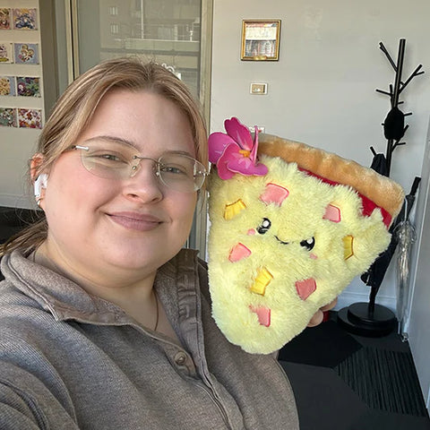 A woman holds the plush Hawaiian pizza slice near her face. It is about the size of her head.