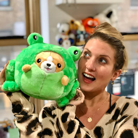 A woman holds the plush corgi in a frog costume near her face. It is slightly larger than her head.