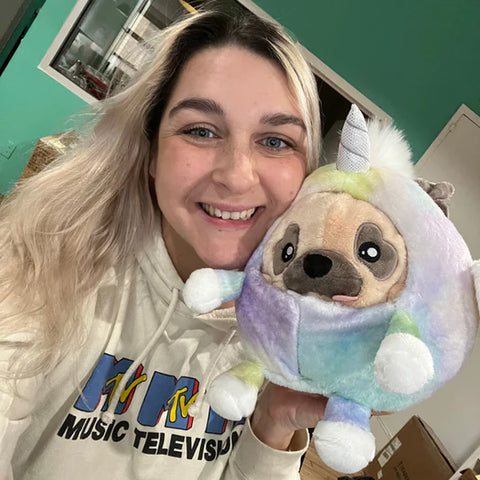 A woman holds the plush pug in a unicorn costume near her face. It is slightly larger than her head.