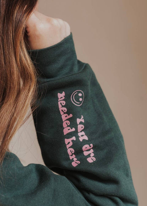 Black hoodie sleeve with a mauve smiley face and text reading "you are needed here"