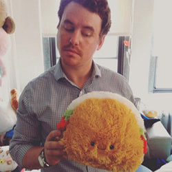 A man holds the plush taco. It is slightly larger than his head.