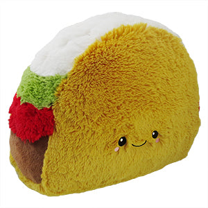 A plush toy taco with a smiling face, made of soft yellow fabric, with a white and green topping on the upper part.