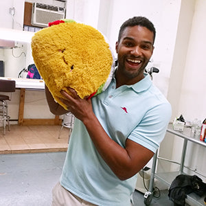 A man holds the plush taco near his head. It is about the size of his torso.