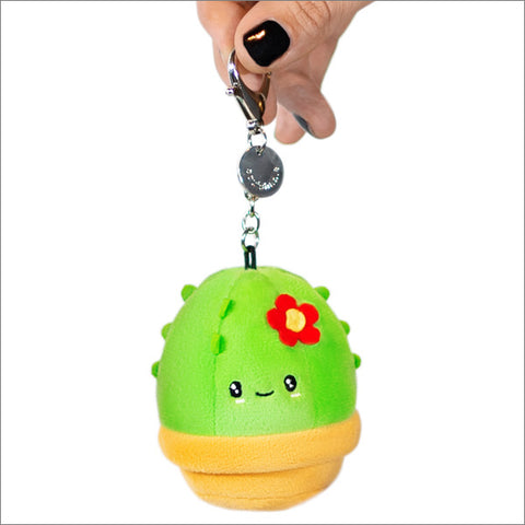 A green cactus-shaped squishy with a happy facial expression, attached to a metal clip which is being held by a hand.