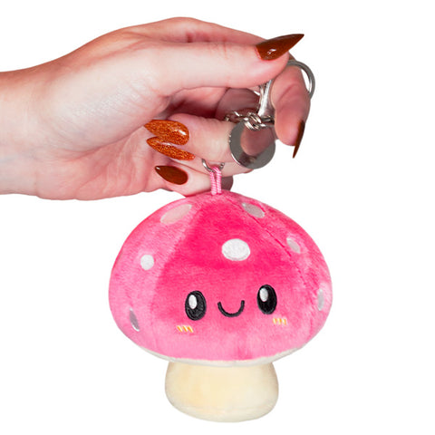A plush toy resembling a pink mushroom with a smiling face, being held by a hand with orange nail polish. The toy has a metal clip on top.