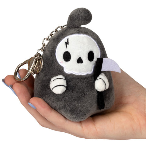A plush toy depicting a reaper character with a scythe, primarily in gray and black colors, held in a person's hand. It has a silver keychain clip attached.
