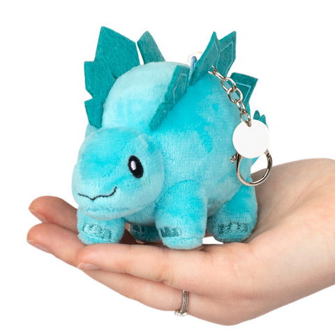 A plush toy resembling a micro Stegosaurus in blue color, being held in a person's hand. It is attached to a silver clip keychain.
