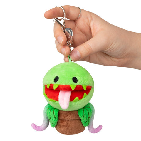 A plush toy resembling a Venus flytrap with a green body, red mouth, and black metal clip, held in a person's hand.