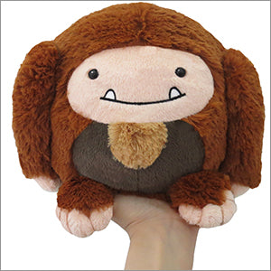 A plush toy resembling a miniature Bigfoot, with brown fur and a friendly facial expression, held in a person's hand.