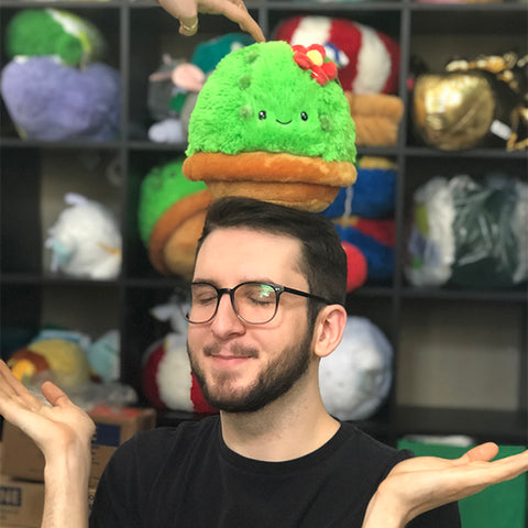 A man balances the plush cactus on his head. It is about the size of his head.