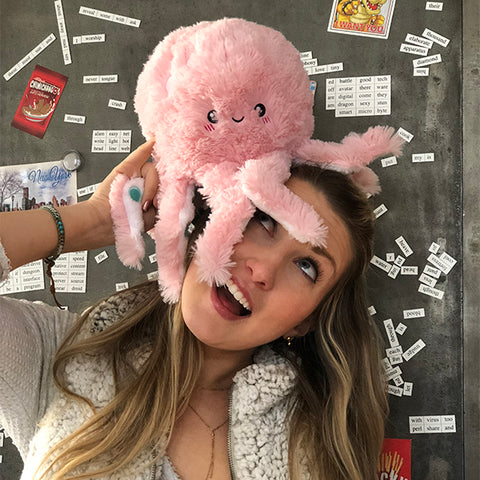A woman holds the plush pink octopus to her head. It is slightly larger than her head.