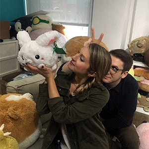 A woman holds up the plush bunny while a man makes bunny ears behind her. The bunny is slightly larger than her head.