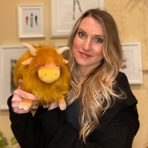 A woman holds the plush highland cow. It is slightly larger than her head.