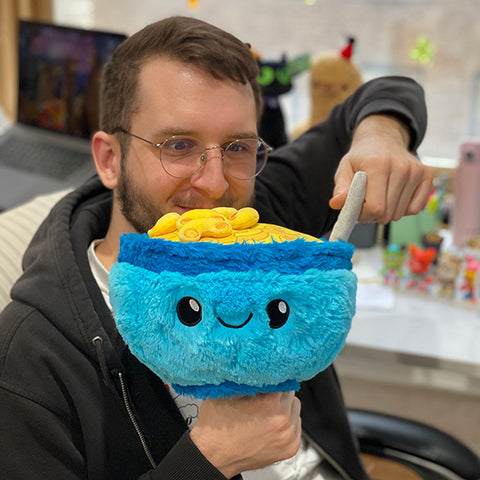 A man holds the plush mac & cheese bowl up to his face. It is slightly larger than his head.