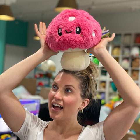 A woman balances the pink plush mushroom on her head. It is slightly larger than her head.