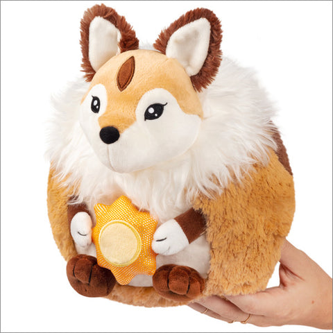 A plush foxlike creature with a white mane, holding a plush sun is held in someone's hand