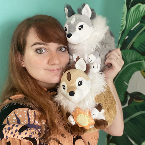 A woman holds two plush toys up to her face. The skoll is slightly larger than her head.