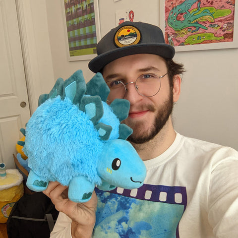A man holds the plush Stegosaurus up to his face. It is slightly larger than his head.