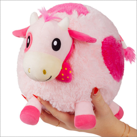 A hand holding a small pink fluffy plush toy shaped like a strawberry cow with a jingle bell around its neck.