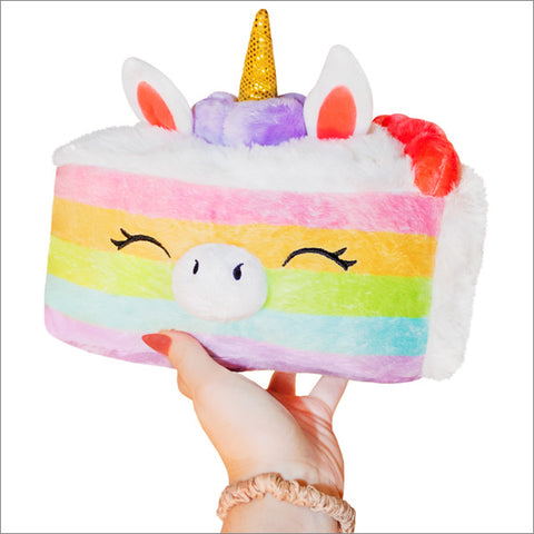 A hand holding a small plush toy resembling a mini unicorn cake with a rainbow cake and face features.