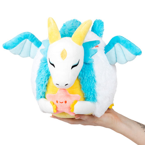 A plush blue and white dragon with yellow horns and belly, holding a plush pink star. It is held in someone's hand.