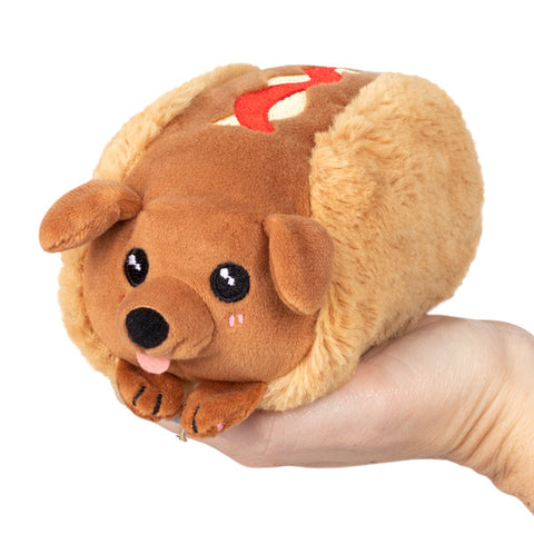 A plush toy resembling a dachshund hot dog, with brown 'meat' and a white 'bread' casing, held in a person's hand.