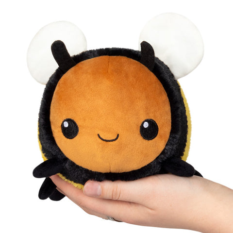 A plush toy designed to look like a bumblebee with a beige body, black and yellow stripes, and white wings. Held on someone's hand.