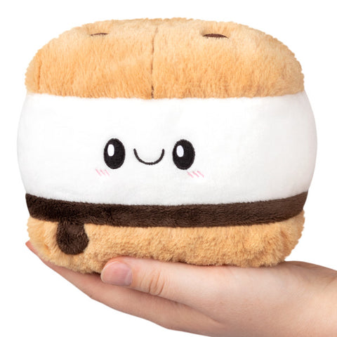A plush toy designed to look like a s'mores sandwich with a smiling face in the center, held in a person's hand.