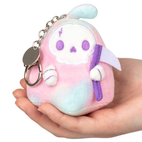 A plush toy in the form of a tie-dye reaper with a pink, blue, and white color scheme, attached with a small keychain, and being held in a person's hand.